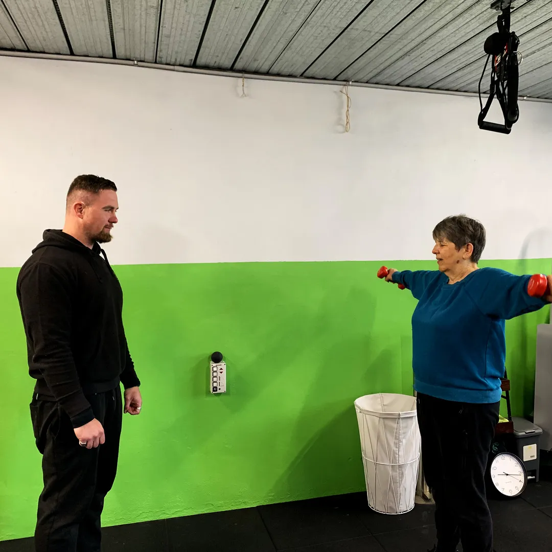 Lachlan standing back, watching a woman raise a light dumbbell to the side.