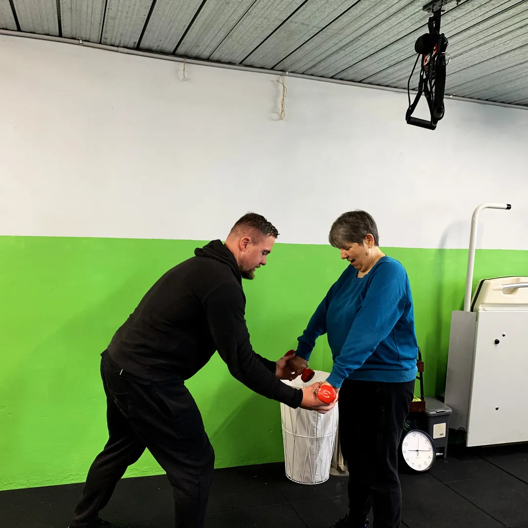 Lachlan guiding a woman's hands on a light dumbbell, against the green wall.